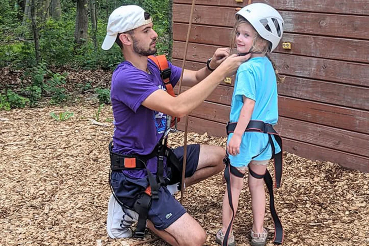 A certified camp counselor straps a helmet onto a young camper as they prepare to use the outdoor climbing wall