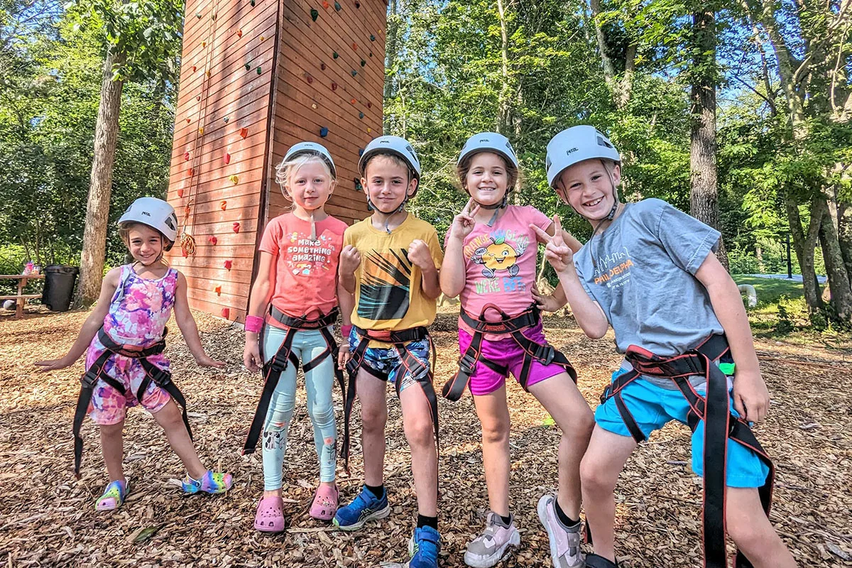 Five campers in helmets and climbing harnesses stand in front of an outdoor climbing tower and pose for the camera
