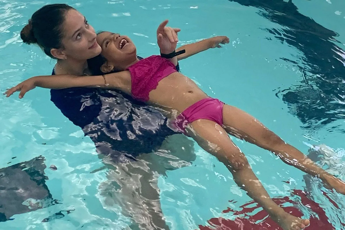 A swim coach supports a young student as the swim student practices floating in the indoor pool