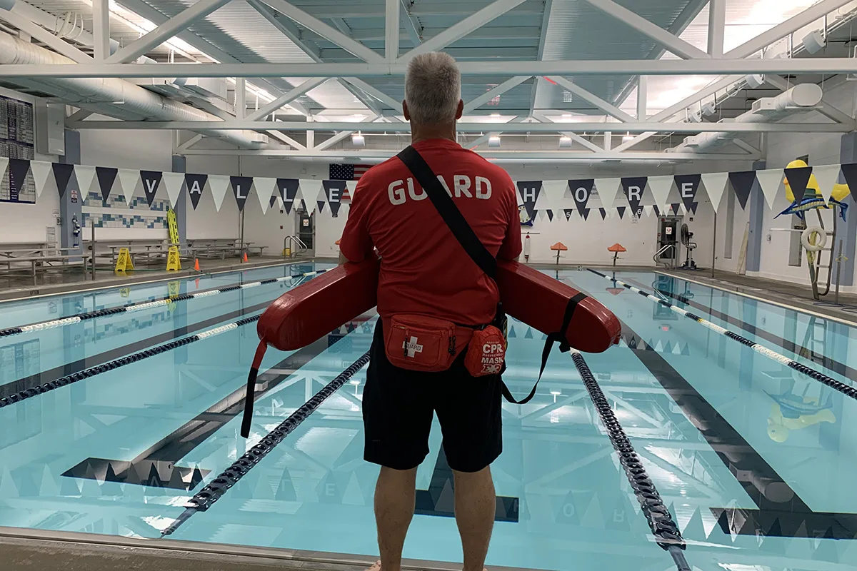 A lifeguard stands equipped with his floatation device and medical kits facing the pool with lanes at Valley Shore YMCA